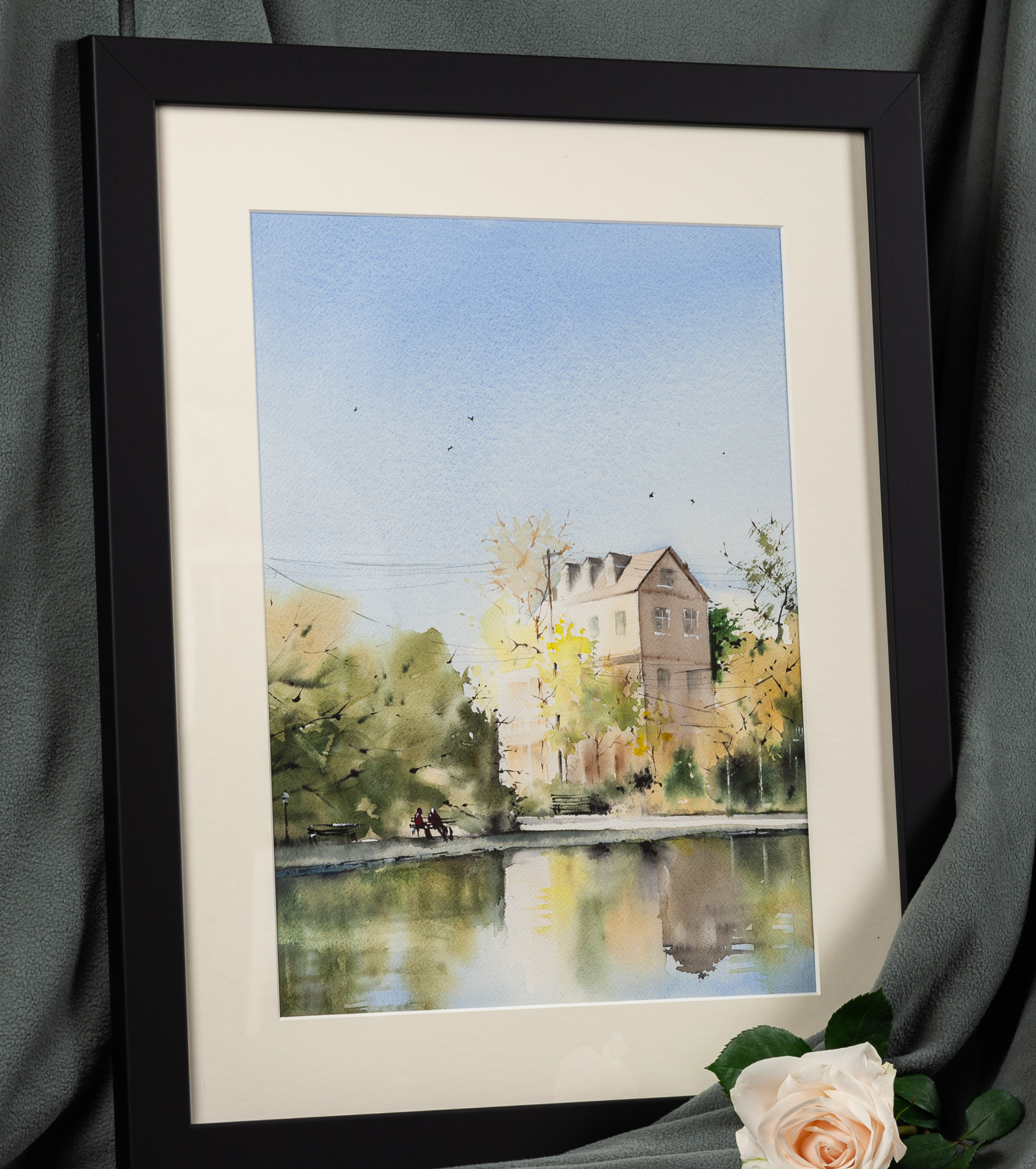 Framed watercolor painting titled Colonial Lake displayed in a black frame with white matting, featuring a Charleston lake scene with trees, a house, and soft reflections in the water.