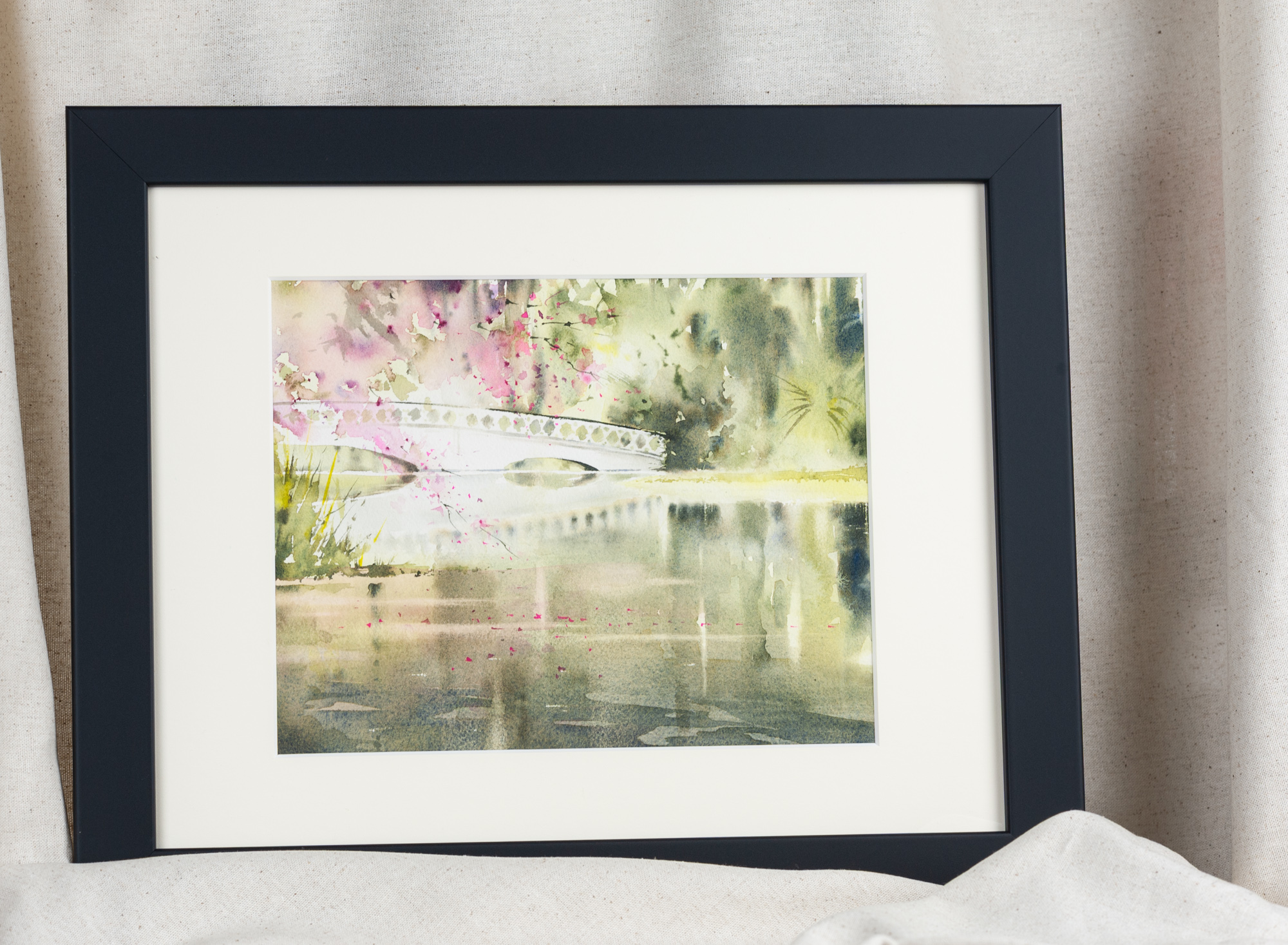 Framed watercolor painting of the iconic white bridge at Magnolia Plantation and Gardens in Charleston, featuring soft pink blossoms and tranquil water reflections, displayed in a black frame with white matting.